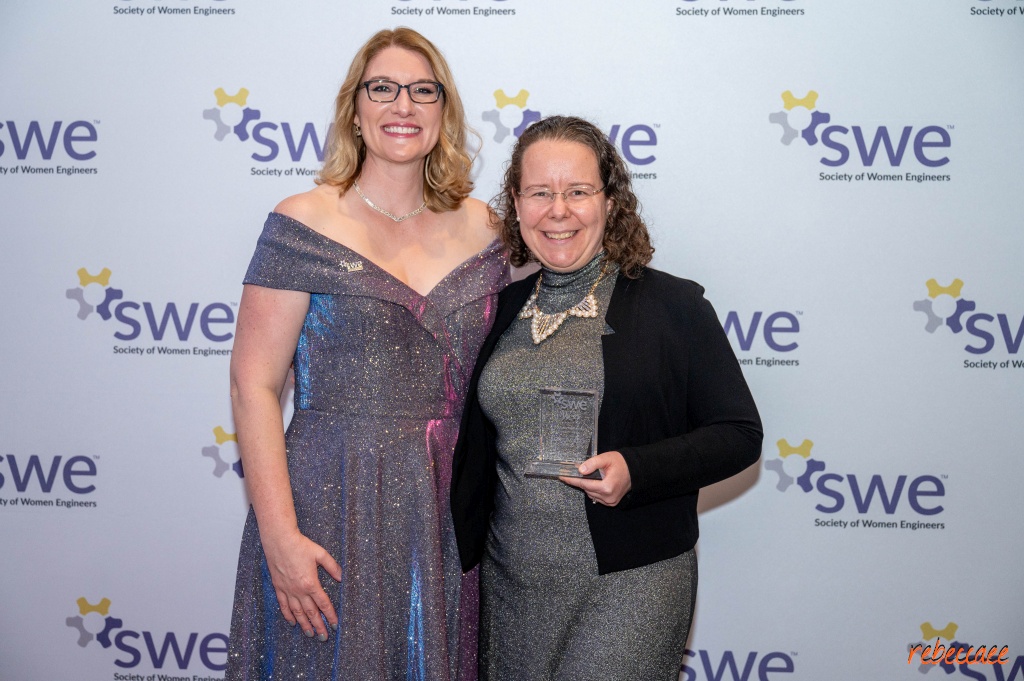 Two women in front of a backdrop with the SWE logo.  (On the left) Alexis is a white woman with shoulder length blonde hair wearing an off-the-shoulder purple sparkling dress.  (On the right) Rebecca is a white woman with curly brown hair wearing a silver sparkling dress and a black jacket.  She is holding an award.