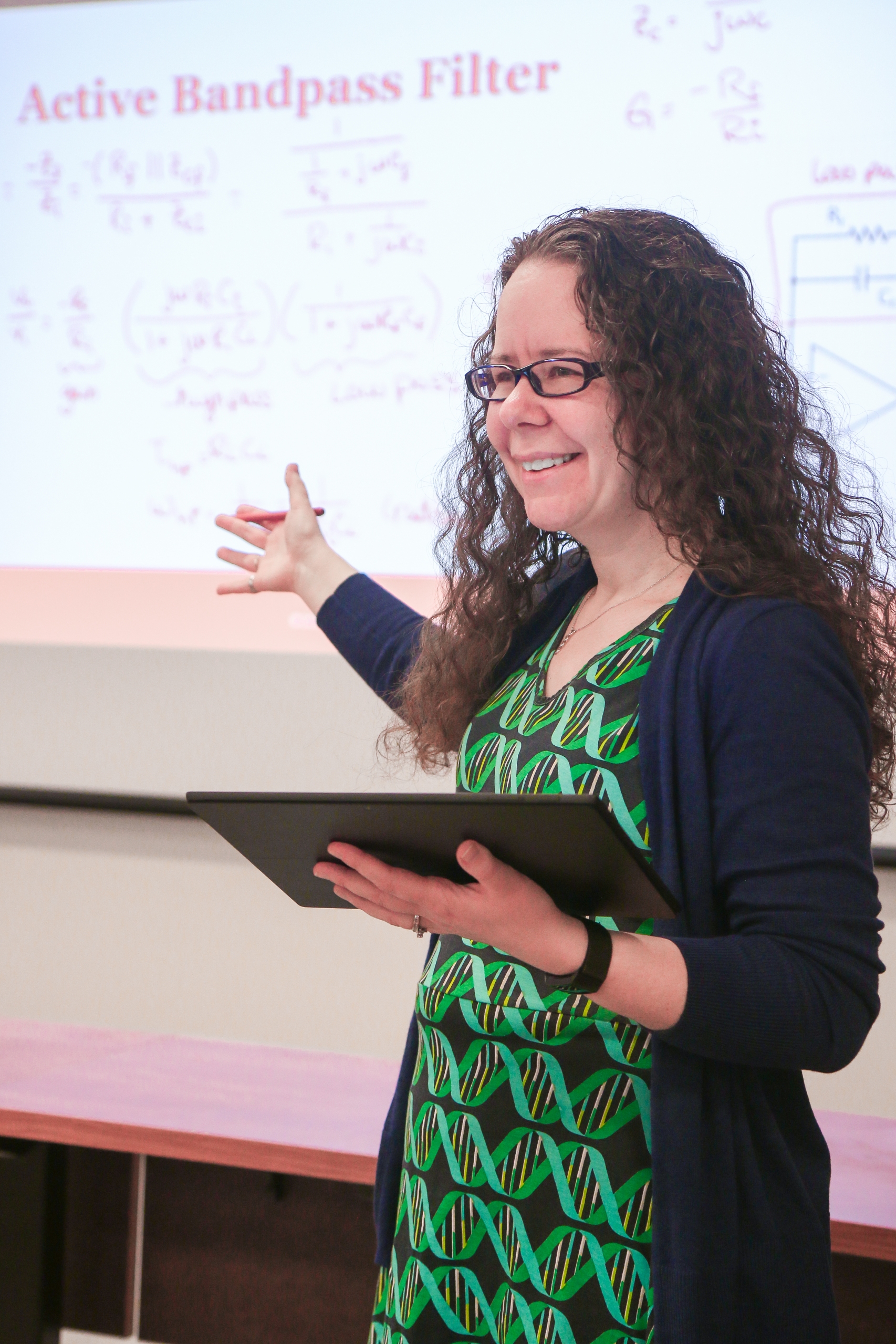 Dr. Reck, a white woman with long curly brown hair and glasses wearing a dress with a dna pattern and cardigan, stands at the front of a classroom with a tablet in one hand and pointing at a screen with the other hand.